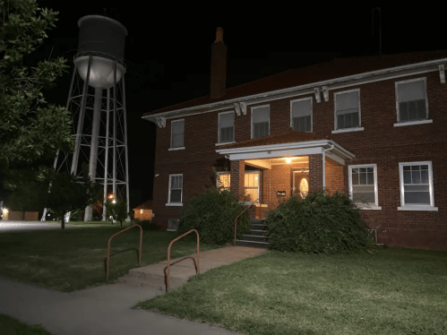 A brick building at night with a water tower in the background, illuminated by warm light at the entrance.