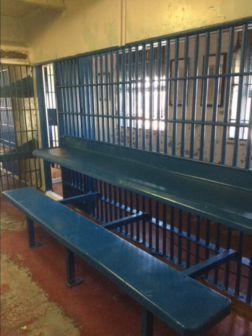Interior of a jail cell featuring blue benches and barred windows, with a worn and aged appearance.