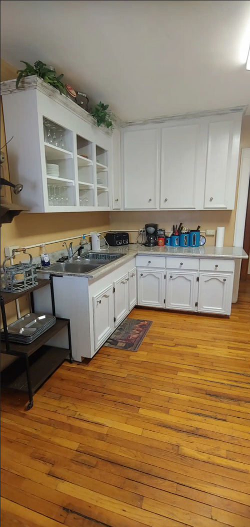 A bright kitchen with white cabinets, a stainless steel sink, and wooden floors, featuring various kitchen appliances.