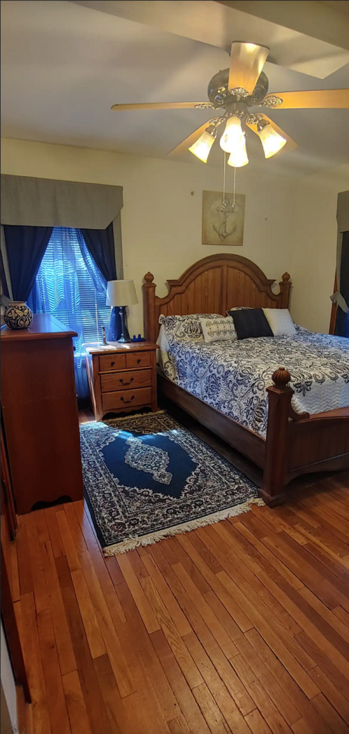 Cozy bedroom with a wooden bed, nightstand, dresser, and a blue patterned rug on hardwood floors. Natural light from window.