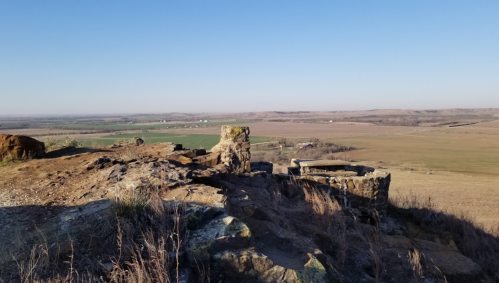 A rocky outcrop overlooks a vast, open landscape with fields and distant hills under a clear blue sky.