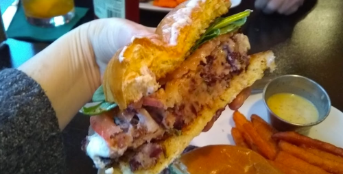 A hand holding a juicy burger with bacon, lettuce, and a side of sweet potato fries on a table.