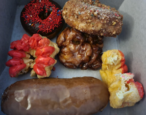 A box of assorted donuts: chocolate with sprinkles, apple fritter, and two frosted varieties, including a long chocolate one.
