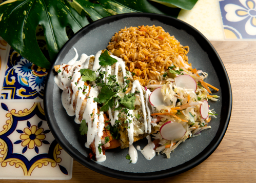 A plate of enchiladas topped with sour cream and cilantro, served with rice and a side of coleslaw.
