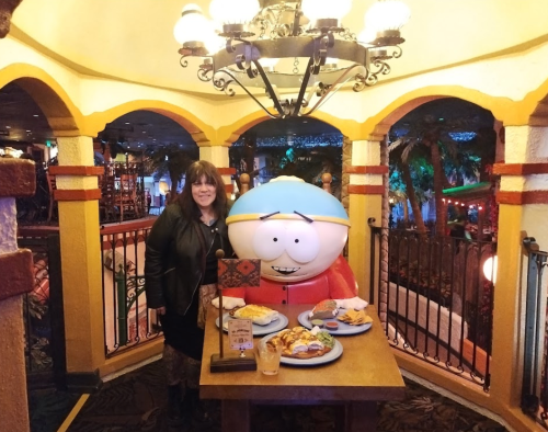 A woman stands next to a large cartoon character statue, with a table of food in front of them in a colorful setting.