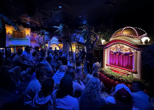 A crowd gathers in a dimly lit theater, watching a puppet show with a colorful stage and vibrant decorations.