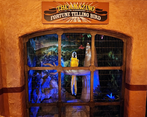 A colorful display featuring a fortune-telling bird behind a barred window, with vibrant lighting and a whimsical sign above.