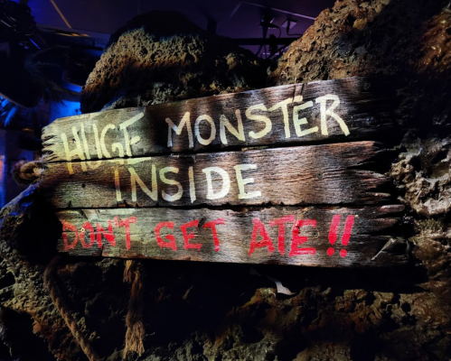 A weathered wooden sign reads "HUGE MONSTER INSIDE DON'T GET ATE!!" against a dark, rocky background.