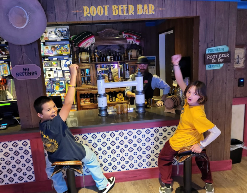 Two children cheer at a bar labeled "Root Beer Bar," while a bartender prepares drinks in the background.