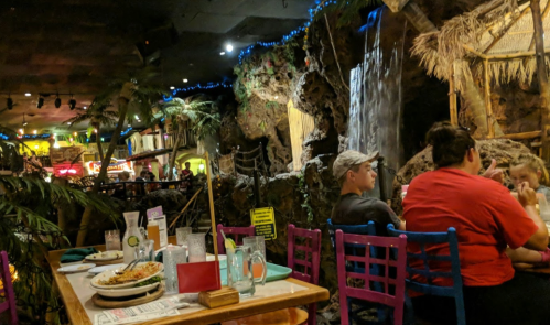 A lively restaurant interior with tropical decor, featuring a waterfall, colorful chairs, and diners enjoying their meals.