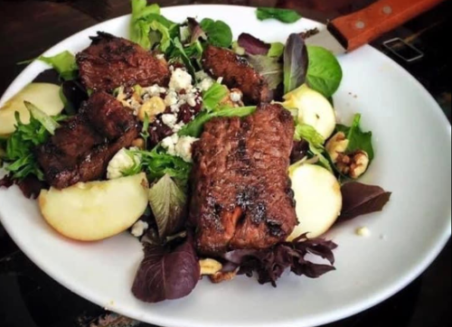 A fresh salad with grilled steak, mixed greens, apple slices, walnuts, and crumbled cheese on a white plate.