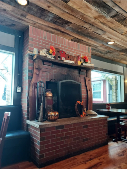 Cozy restaurant interior featuring a brick fireplace adorned with seasonal decorations and wooden beams overhead.
