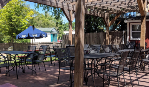 Outdoor seating area with metal tables and chairs under a wooden pergola, surrounded by greenery and a blue umbrella.