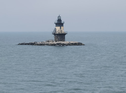 A black and white lighthouse stands on a rocky island in the ocean, surrounded by calm waters under a gray sky.