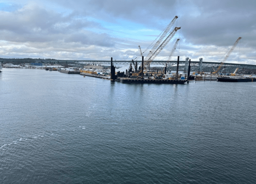 A view of a harbor with cranes, boats, and a bridge under a cloudy sky. Water reflects the surroundings.