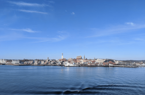 A scenic view of a waterfront city under a clear blue sky, with buildings and boats reflected in the calm water.