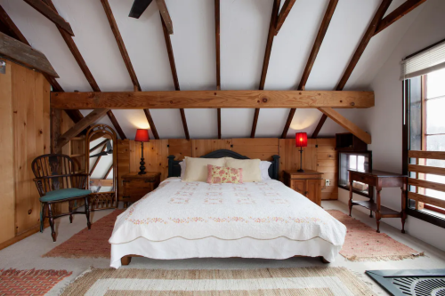 Cozy attic bedroom with wooden beams, a large bed, red lamps, and rustic decor. Natural light from a window.