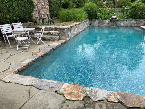 A clear blue swimming pool surrounded by stone walls and a small table with chairs nearby in a lush garden setting.