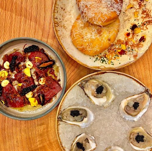 A wooden table with three plates: oysters on ice, a colorful tuna dish, and fried dough balls topped with cheese.