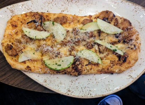 A flatbread pizza topped with sliced apples, cheese, and herbs on a decorative plate.