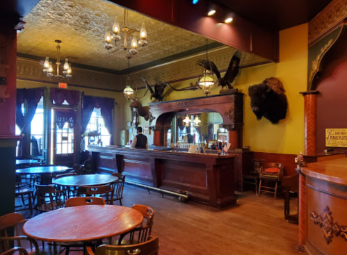 A vintage bar interior with wooden tables, chandeliers, and mounted animal heads on the walls.