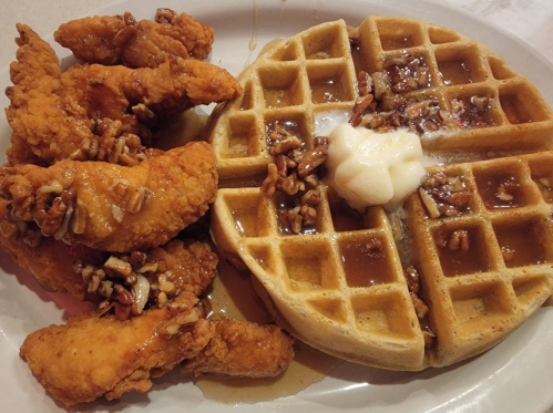 A plate featuring crispy fried chicken tenders and a golden waffle topped with pecans and syrup.