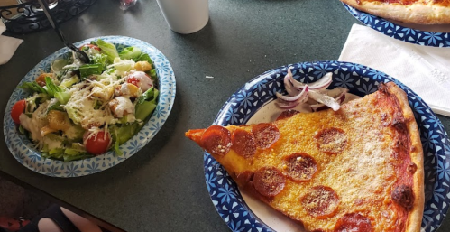 A slice of pepperoni pizza on a plate next to a salad with greens, tomatoes, and croutons on a table.