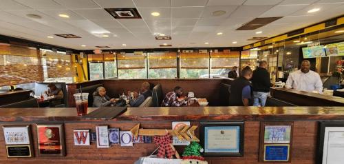 A cozy restaurant interior with booths, patrons dining, and a counter with staff serving food. Holiday decorations visible.