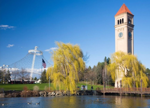 A scenic view of a clock tower and a large Ferris wheel by a lake, with willow trees and blue skies in the background.