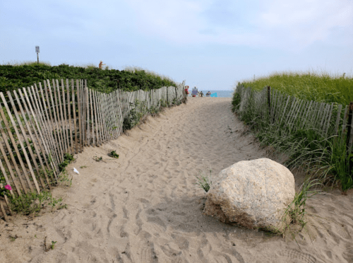 A sandy path leads to the beach, flanked by wooden fences and greenery, with a large rock in the foreground.