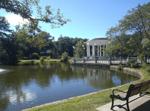 A serene park scene featuring a gazebo by a calm pond, surrounded by trees and a bench in the foreground.