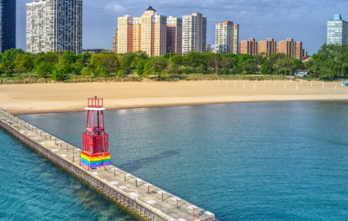A colorful lighthouse stands on a pier by a calm lake, with sandy beach and city buildings in the background.