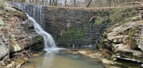 A serene waterfall cascading over a stone wall into a calm stream, surrounded by rocky terrain and trees.