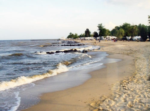 A sandy beach with gentle waves, rocky shoreline, and campers in the background under a clear sky.