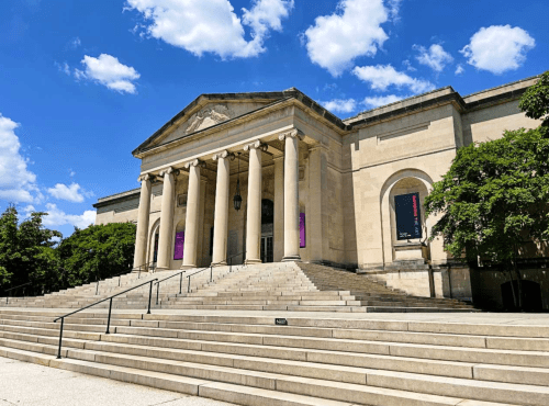 A grand museum entrance with tall columns, wide steps, and a blue sky with fluffy clouds above.