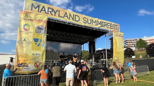 A crowd watches a band perform on stage at Maryland Summerfest, with a large banner in the background.