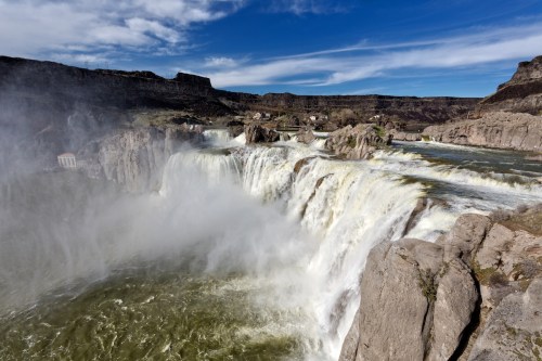 A stunning waterfall cascades over rocky cliffs, surrounded by mist and a blue sky with wispy clouds.