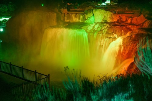 A vibrant waterfall illuminated in green and orange lights, surrounded by lush vegetation and a viewing platform.