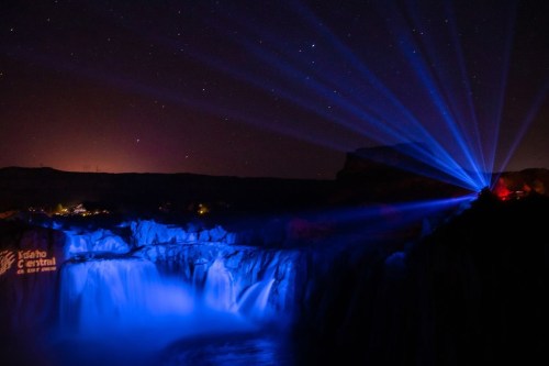 A nighttime view of a waterfall illuminated by blue lights, with stars visible in the sky above.