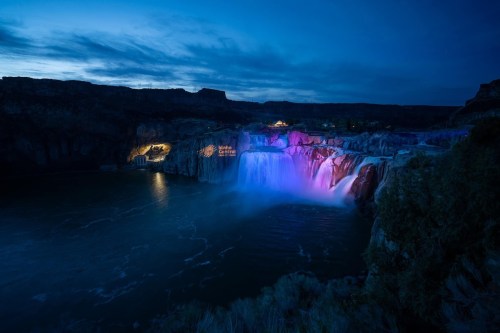 A colorful waterfall illuminated at night, surrounded by rocky cliffs and a calm river.
