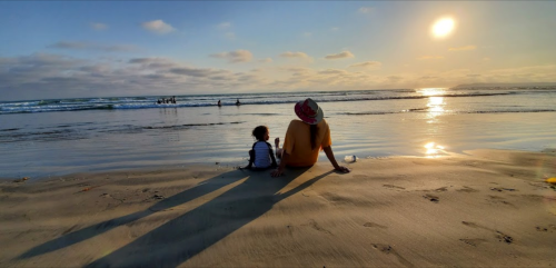 A person and a child sit on the beach, watching the sunset over the ocean, with gentle waves in the background.