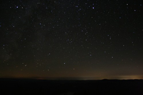 A clear night sky filled with countless stars, with a faint glow on the horizon and dark silhouettes of distant hills.