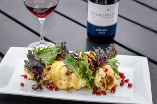 A plate of roasted cauliflower topped with greens and pomegranate seeds, accompanied by a glass of red wine.