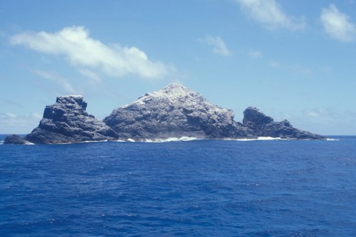 A rocky island rises from the blue ocean under a clear sky with a few clouds. Waves gently lap at its base.