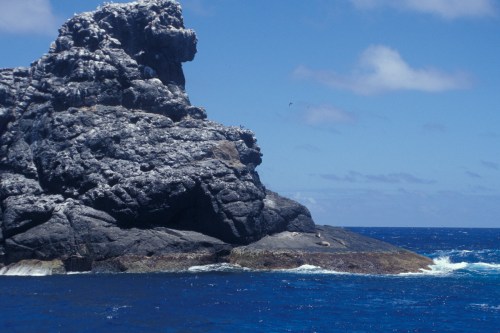A rugged rock formation rises from the ocean under a clear blue sky, with gentle waves lapping at its base.