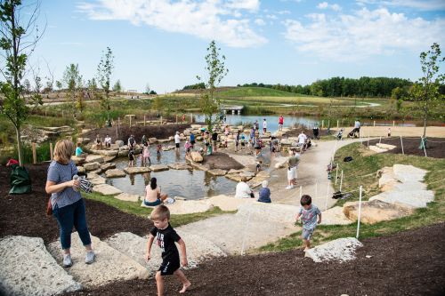 A lively park scene with children and adults playing by a pond, surrounded by greenery and walking paths.