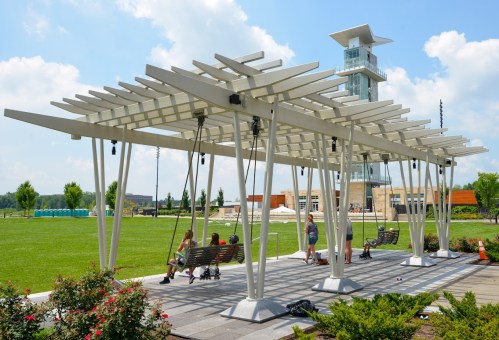 A modern pavilion with swings, surrounded by greenery and a tall building in the background under a blue sky.