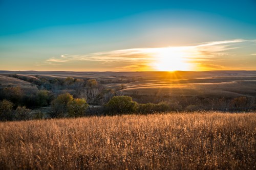 A stunning sunset over the rolling Flint Hills of Kansas, with golden light illuminating the tallgrass prairie and soft clouds glowing in warm orange and pink hues across the horizon.