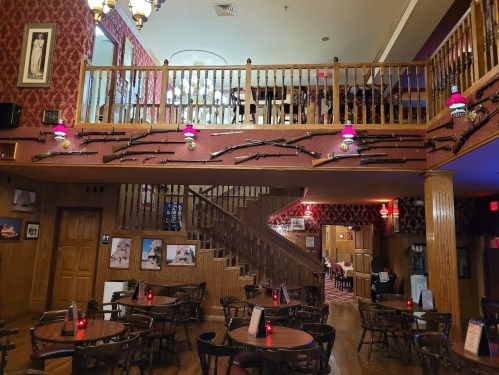 Interior of a rustic restaurant with wooden decor, tables, and vintage rifles displayed on the walls.