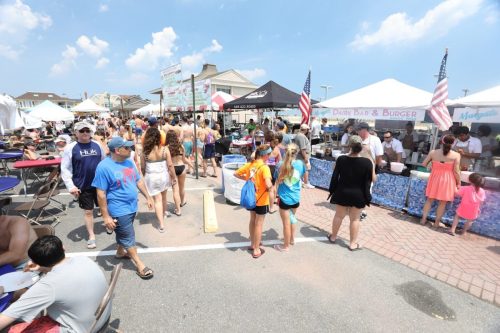 A bustling outdoor market with food stalls, people walking, and colorful tents under a clear blue sky.